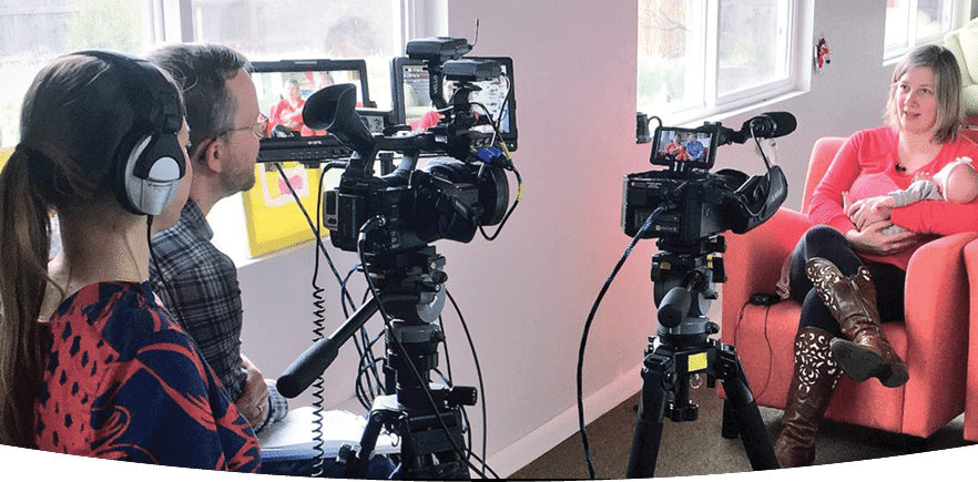 Looking over the shoulders of 2 people sitting in behind cameras that are capturing the interview of a woman holding a baby sitting in an arm chair