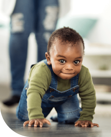 Toddler wearing a green long sleeve shirt and overalls is crawling across the floor with out of focused legs of an adult seen in the background