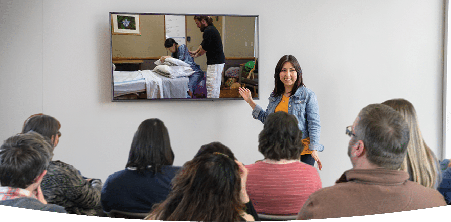 A teacher in front of a classroom of adults pointing to a video playing on the screen