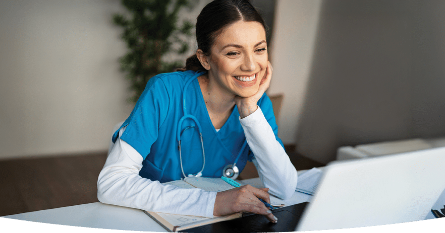 Smiling nurse in scrubs sitting at a desk with a notebook in front of her and looking at a laptop 
