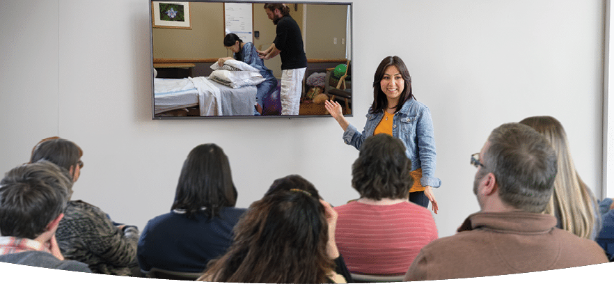A teacher in front of a classroom of adults pointing to a video playing on the screen
